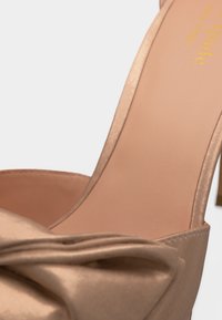 Κοντινό πλάνο ενός μπεζ παπουτσιού με ψηλό τακούνι και μεγάλο σατέν φιόγκο στη μπροστινή λουρίδα.