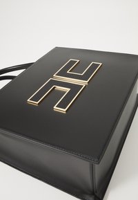 Black leather handbag with a smooth texture, featuring a large gold and textured black emblem shaped like an "H" on the front.