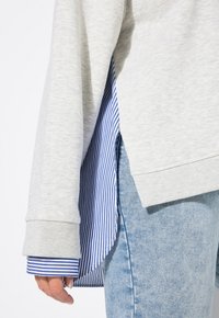 Sudadera gris con puños acanalados sobre una camisa a rayas azul y blanca, con un dobladillo curvado y diseño en capas, combinada con jeans de mezclilla.