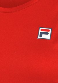 Camiseta de algodón roja con cuello redondo, que presenta un logo rectangular de Fila en azul, rojo y blanco en el lado izquierdo.