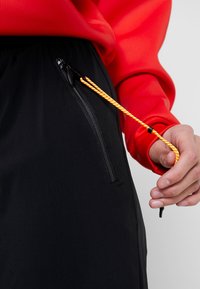 Černé sportovní šortky mají zipovou kapsu a oranžovou šňůrku na stažení. Pás je elastický pro větší pohodlí. Červená mikina je částečně viditelná.