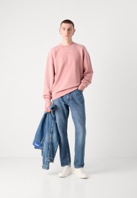 Sudadera blanca de gran tamaño, jeans de mezclilla azules de corte recto y zapatillas blancas. Sosteniendo una chaqueta de mezclilla con detalles azul.