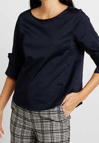 Mörkblå blus med trekvartsärmar gjord av mjukt tyg. Har en rund halsringning och diskreta sid sömmar, kombinerad med rutiga byxor.