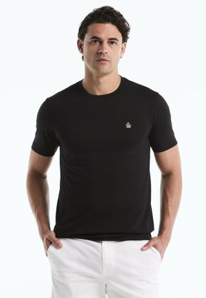 Mann mit dunklem Haar, trägt ein schwarzes T-Shirt mit kurzen Ärmeln und kleinem Logo auf der Brust sowie weiße Hosen, die Hände in den Taschen.