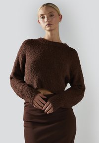 Kobieta z blond włosami, ubrana w teksturowany brązowy sweter do pasa i dopasowaną spódnicę, stojąca na tle jasnego, jednolitego tła.