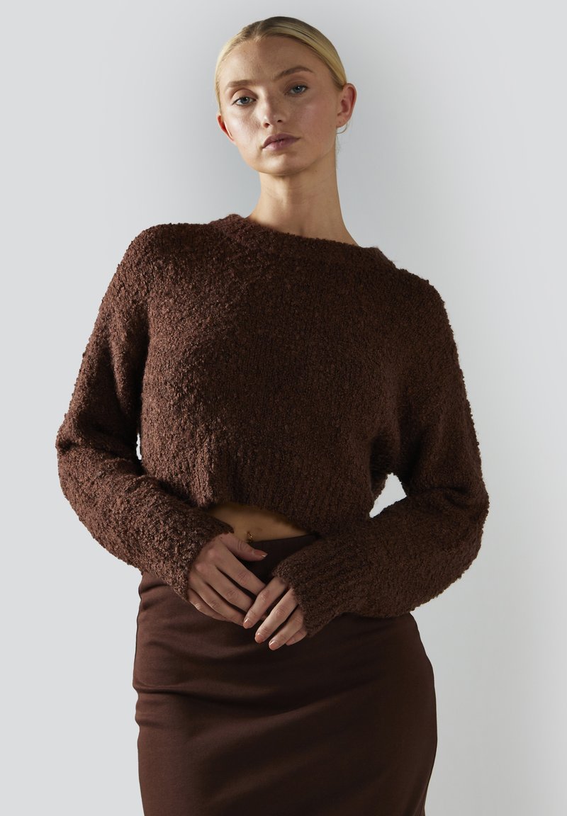 Kobieta z blond włosami, ubrana w teksturowany brązowy sweter do pasa i dopasowaną spódnicę, stojąca na tle jasnego, jednolitego tła.