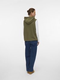 Πράσινο φλις γιλέκο με κουκούλα, συνδυασμένο με λευκό μακρυμάνικο πουκάμισο, μπλε τζιν και καφέ παπούτσια. Ορατή λεπτομέρεια στο πίσω μέρος και άνετη εφαρμογή.