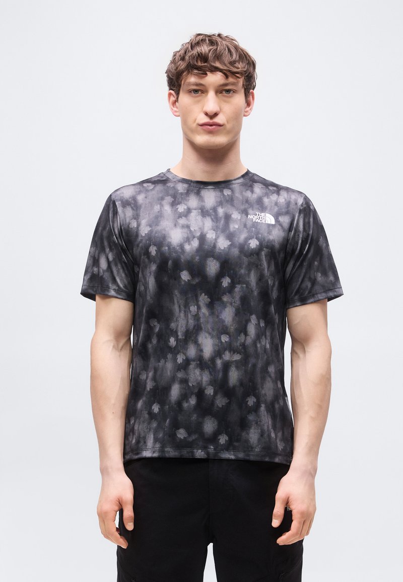 Νέος άνδρας με κοντά, σγουρά μαλλιά, φοράει κοντομάνικη μπλούζα The North Face σε μαύρο και γκρι με σχέδιο, στέκεται μπροστά σε ένα φωτεινό φόντο.