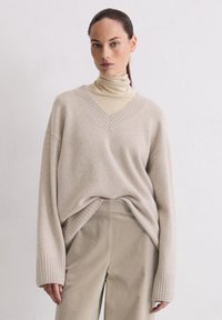 Beige oversized trui met V-hals en geribde manchetten, gelaagd over een lichte coltrui. Zachte textuur; ontspannen pasvorm met een ruime silhouette.
