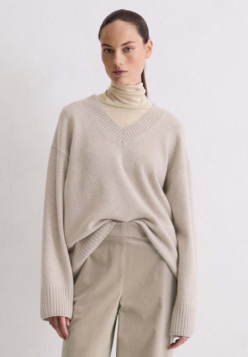 Beige oversized trui met V-hals en geribde manchetten, gelaagd over een lichte coltrui. Zachte textuur; ontspannen pasvorm met een ruime silhouette.