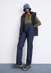 Jachetă puffer neagră peste un tricou cu mânecă lungă în nuanță de măslin, combinată cu blugi închiși la culoare, cu talie înaltă, și adidași gri cu accente verzi.