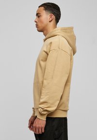 Felpa beige con cappuccio regolabile, tasca laterale e polsini a coste. Materiale morbido, vestibilità rilassata, texture liscia e design semplice.