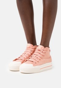 Rosa canvas hög-top sneakers med vita gummisulor och snören, med varumärke på tungan och en texturerad yta.