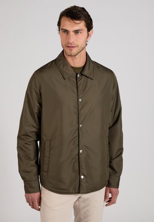 Mężczyzna z krótkimi ciemnymi włosami, ubrany w zapinaną na guziki lekką oliwkowo-zieloną kurtkę i beżowe spodnie na jednolitym tle.