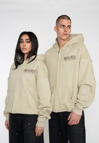 Des sweatshirts zippés beiges en tissu doux, avec de grandes poches avant et un design graphique sur la poitrine gauche. Des jeans foncés contrastent avec les sweatshirts.