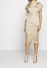 Abito beige fino al ginocchio con cintura in vita, maniche corte e tasche ispirate al design militare. Abbinato a tacchi bianchi con cinturini.