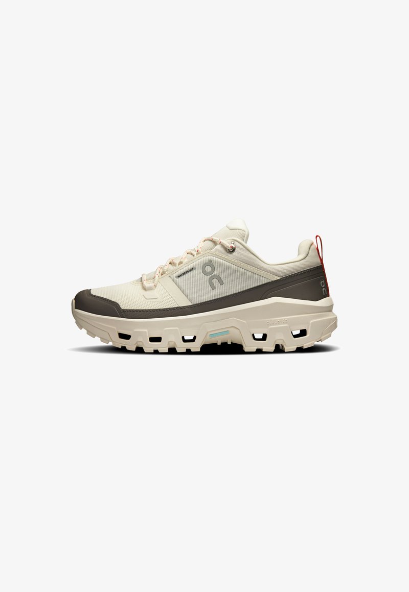 Leichte beige Sneaker mit einem Obermaterial aus strukturiertem Stoff, dunkelbraunen Akzenten, Gummisohle und einer roten Schlaufe am Fersenbereich.
