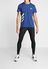 Maglietta atletica blu a maniche corte con design a strisce bianche, abbinata a leggings neri aderenti e scarpe da ginnastica bianche con dettagli blu.