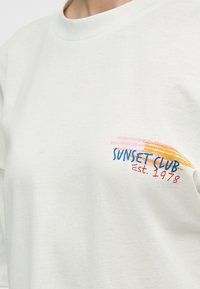 Λευκό μπλουζάκι με το κείμενο "SUNSET CLUB Est. 1978" και σχέδιο από ροζ και πορτοκαλί πινελιές στη αριστερή πλευρά του στήθους.