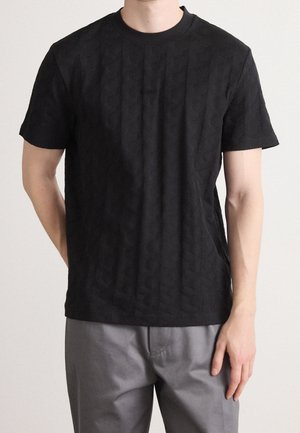 Mężczyzna w czarnej, teksturowanej koszulce z krótkim rękawem i szarych spodniach stoi na tle jednolitego tła.