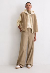 Beige Fleeceweste mit Reißverschluss vorne über einem cremefarbenen Hoodie, kombiniert mit langen beigefarbenen Hosen und weißen Plateausneakern mit roten Akzenten.