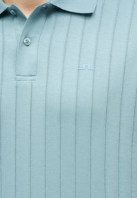 Polo bleu clair côtelé avec un logo brodé discret sur la poitrine gauche, avec un col et deux boutons près de l'encolure.