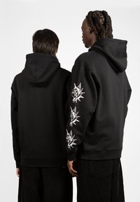 Μαύρες φούτερ από μείγμα βαμβακιού διαθέτουν καπιτονέ κουκούλα και λευκές γραφικές εκτυπώσεις κεφαλών λύκων στα μανίκια. Η πίσω πλευρ�ά δείχνει δύο χρήστες.