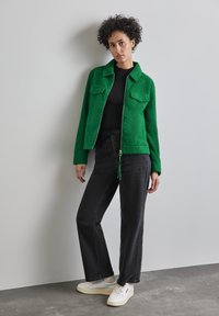 Grön texturerad jacka med två framfickor, dragkedjestängning och dragsko. Kombinerad med svarta byxor och vita sneakers på en grå bakgrund.