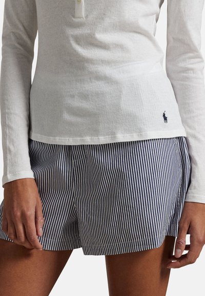 Vrouw die een wit poloshirt met lange mouwen en blauw-witte verticale gestreepte korte broek draagt, staand met ontspannen handen langs haar lichaam.