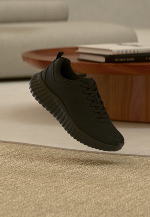 Chaussure de sport noire avec semelle épaisse texturée flottant au-dessus d'un tapis beige devant une table basse en bois avec des livres empilés.