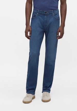 Homme portant un jean bleu et des baskets beiges, debout avec les bras détendus le long du corps sur un fond uni.