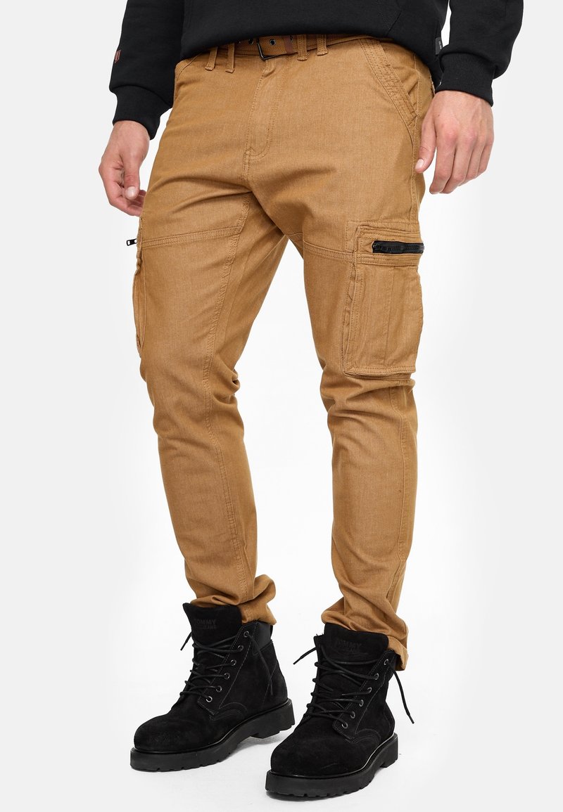 Homme portant un pantalon cargo beige avec plusieurs poches et des bottes noires à lacets, debout les mains détendues le long du corps.