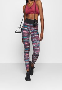 Bordeaux sportbeha met logo, kleurrijke leggings met een patroon in blauw, roze en wit, gecombineerd met zwarte sneakers en een kleine zwarte tas.