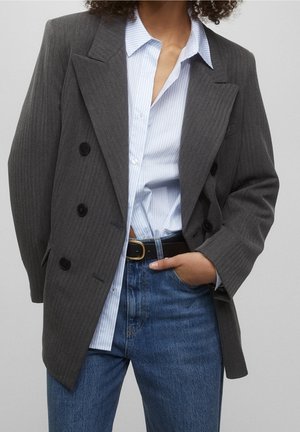 Person wearing a gray double-breasted blazer over a blue and white striped shirt, tucked into high-waisted blue jeans with a black belt.