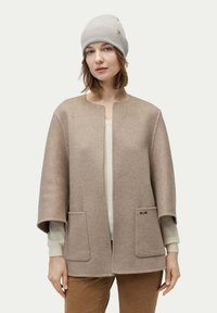 Beige ullblandad rock med rund halsringning och två framfickor; matchad med en ljusfärgad tröja och en ribbad grå mössa.