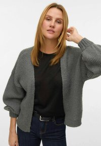 Szara, dziergana kardigan z luźnym krojem, szerokimi rękawami i otwartym przodem, noszony na czarną bluzkę, w połączeniu z ciemnymi dżinsami.