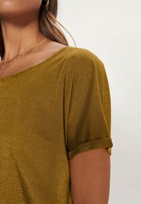 Zlatě zbarvený top s krátkým rukávem a širokým výstřihem, vyrobený z texturované, třpytivé látky. Jsou viditelné stočené rukávy a jemný fall.