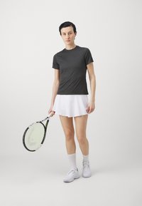 Camiseta atlética de manga corta en gris oscuro combinada con una falda de tenis blanca con pliegues, sosteniendo una raqueta de tenis, zapatillas blancas y calcetines de caña baja.