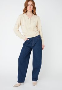 Beige neulepusero, jossa on kaulus ja koristeellisia yksityiskohtia. Yhdistetty leveälahkeisten sinisten denimhousujen ja vaaleansävyisten kärjellisten kenkien kanssa.