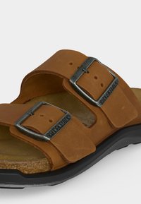 Καφέ δερμάτινο σανδάλι με δύο ρυθμιζόμενες μεταλλικές αγκράφες με χαρακτήρα "BIRKENSTOCK" σε ένα διαμορφωμένο φελλό πάτο.