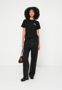 T-shirt preta de algodão com mangas curtas e um detalhe de bordado floral no peito, combinada com jeans pretos de perna larga e uma pequena mala bordô.