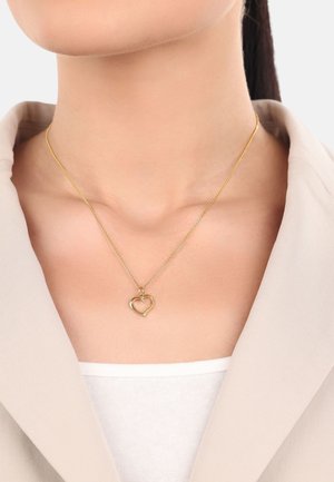 Collana pendente d'oro con un ciondolo a forma di cuore, su una delicata catena, indossata sopra una maglia di colore chiaro con colletto a blazer.