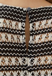Tissu brodé noir et blanc avec motif floral, présentant un design avec une ouverture en forme de clé à l'arrière et une fermeture par un unique bouton noir.