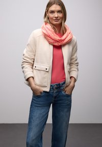 Veste en polaire blanche avec deux poches avant, pull corail en dessous, et une écharpe infinie rose à motif. Un jean bleu à jambes larges complète la tenue.