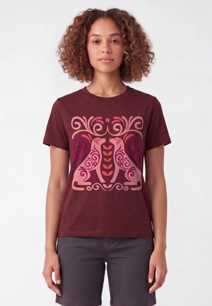 Femme aux cheveux bouclés portant un t-shirt marron avec un motif symétrique rose et violet représentant deux chiens stylisés qui se font face.
