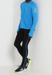 Modrá sportovní tričko s dlouhým rukávem se třemi bílými pruhy na rukávu, doplněné černými legínami s modrým pruhy na boku a černými teniskami.