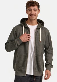 Sudadera con cremallera de color verde oscuro con cordones crema, tela texturizada, bolsillos frontales y capucha interior en contraste. Llevada sobre una camiseta blanca.