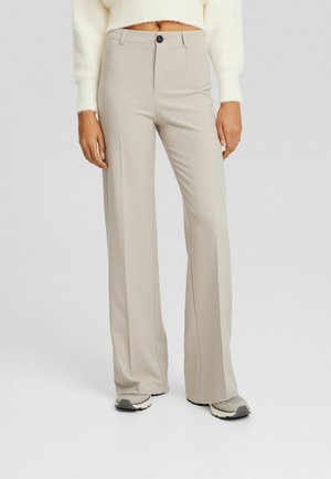 Beige hoge taille wijdere broek met knoop aan de voorkant, gedragen met een witte cropped trui en beige sneakers op een lichtgrijze achtergrond.