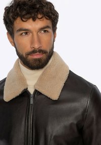 Bruine leren jas met een beige schapenvachtkraag, ritssluiting en een gladde textuur. Draagt over een lichtgekleurde coltrui.