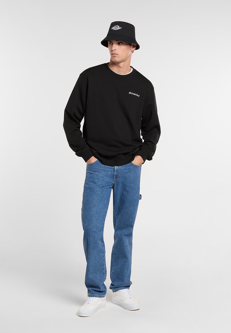 Zwarte sweatshirt met logo, blauwe spijkerbroek, witte sneakers en een zwarte bucket hat. Eenvoudig, casual ontwerp met een losse pasvorm.